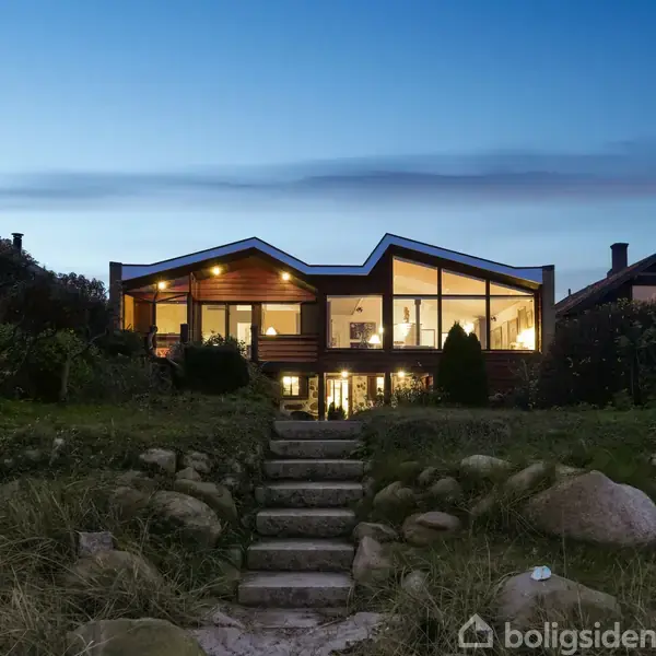 Et moderne hus med store glaspartier lyser op om aftenen. Det står på en frodig, stenet plæne med trapper i forgrunden. Mørk himmel og nabohuse i baggrunden.