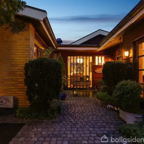 Hus med oplyste vinduer om aftenen, skaber en hyggelig stemning. Flisebelagt gårdhave omgivet af tætte buske. Indendørs lys ses gennem dørene, mens himlen er mørkeblå.