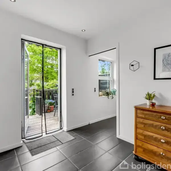 En entré med sort klinkegulv fører ud til en balkon omgivet af grønt løv. En trækommode med en lampe og plante står til højre, en indrammet tegning hænger over den.