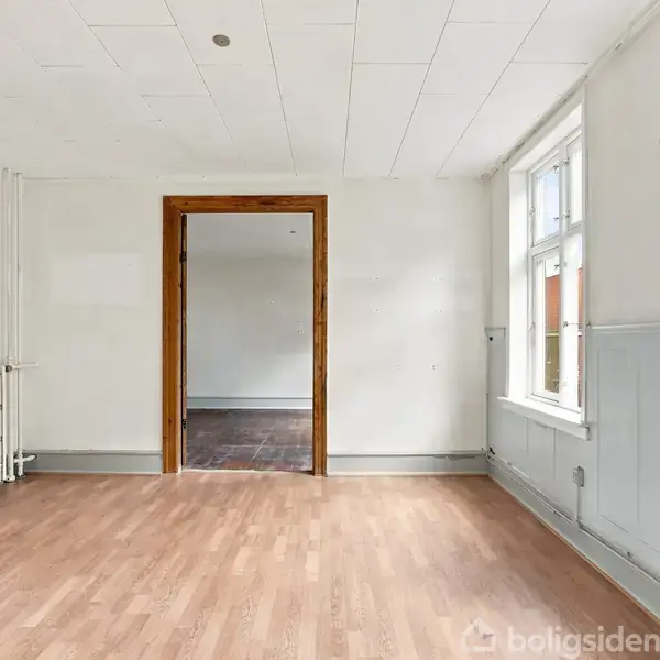 Et rummeligt værelse med trægulv og hvide vægge har en åbning til et tilstødende rum. Store vinduer på højre side tillader naturligt lysindfald. En radiator står til venstre.