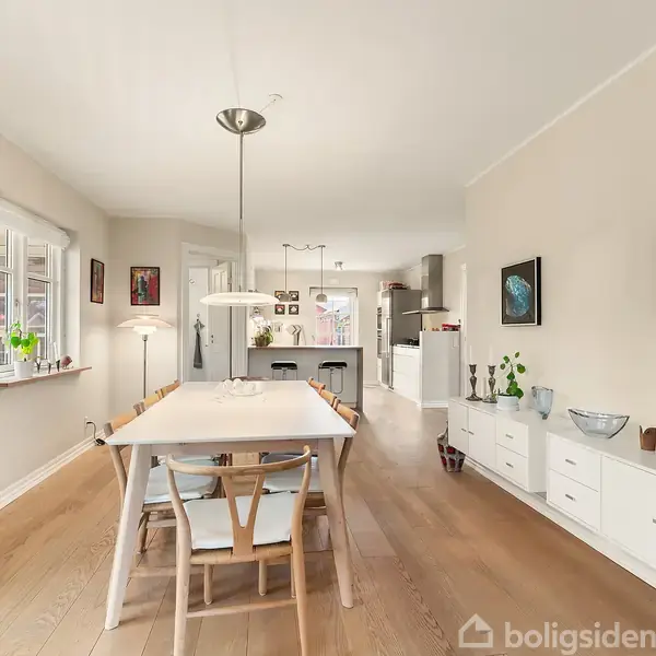 Spisestue med et langt hvidt bord og seks stole, omgivet af moderne kunst på væggene. Skabe langs væggen til højre, lys vedhængslampe, og køkken i baggrunden.