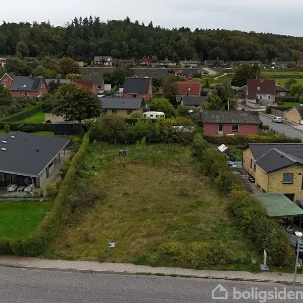 Åben græsmark omgivet af parcelhuse. Græsset er vildt og ujævnt. I baggrunden er der flere huse med haver, og en campingvogn kan ses blandt træerne.