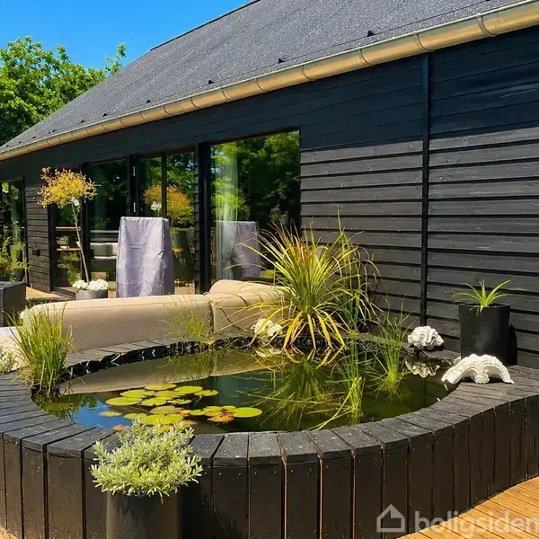 Sort træhus med stor terrasse, hvor en rund dam med planter og vandliljer er centralt placeret. Sofaer og dekorationer omringer dammen. Skyfri blå himmel og træer i baggrunden.