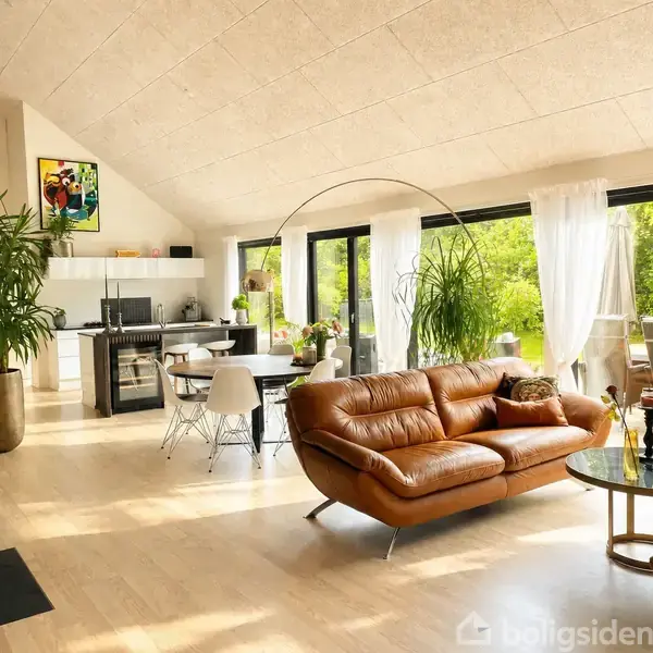 Brun lædersofa står i moderne stue med store vinduer og forkromet sofabord. Til venstre er der en åben køkkensektion med hvide møbler og grønne planter.