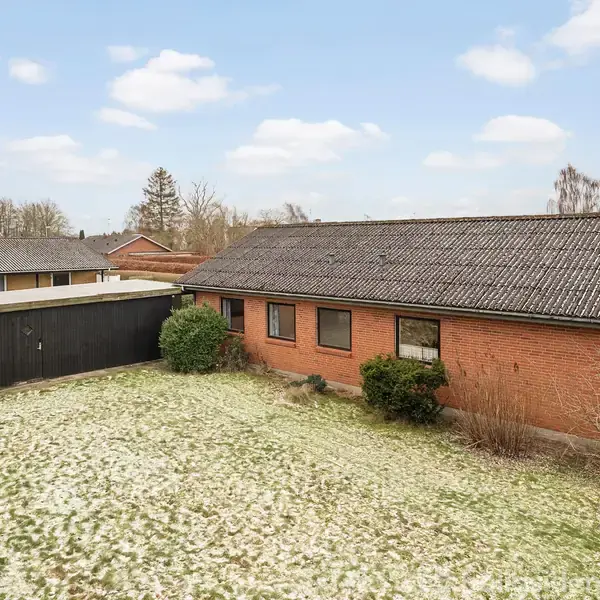 En enkel, rød murstensbygning står stille på en let snedækket græsplæne ved siden af en sort garagebygning. I baggrunden ses flere træer og blå himmel med skyer.