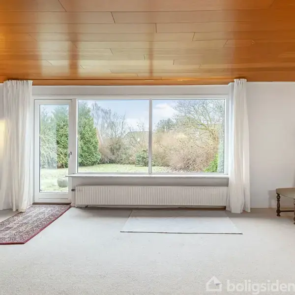 Et rummeligt rum med lyst tæppe og træloft. To stole står nær væggen, mens en stor glasdør med vindue viser en have udenfor. To lamper hænger fra loftet.