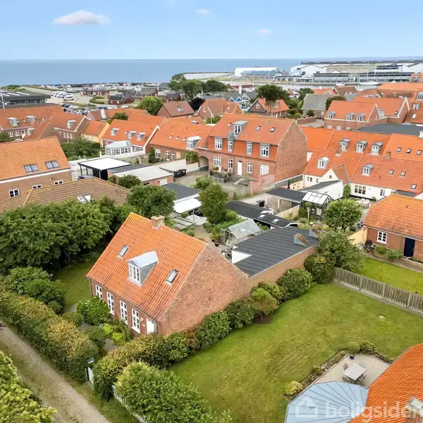 Røde tage pryder boligkvarteret, mens husene stille står langs veje. Haver og træer omgiver hjemmene, der ligger tæt ved en kystlinje. I baggrunden kan man se havet og en by.