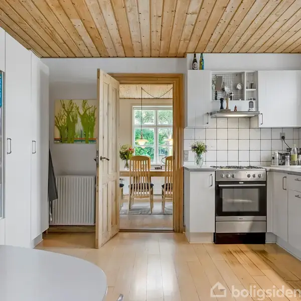 Køkken med hvide skabe og en ovn, der åbner til en spisestue gennem en trædør. Trægulv og træloft skaber en hyggelig atmosfære. Bøger i en reol ved siden af døren.