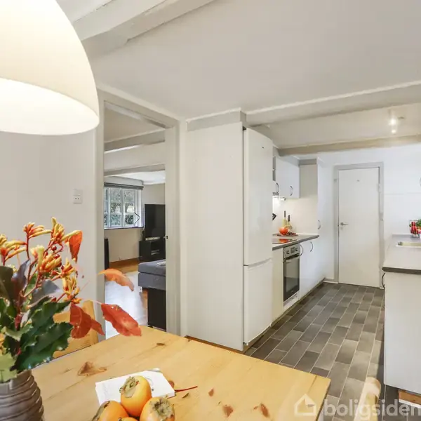 Et bord med en vase med blomster og frugter står i et køkken. Rummet har hvide skabe, en åbning til stue og træbjælker i loftet.