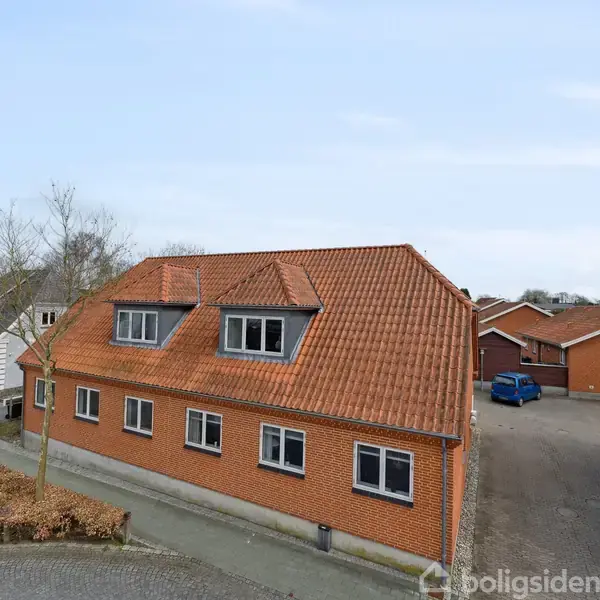 En rødstensbygning med rødt tegltag står stille på et hjørne ved en vej omgivet af andre huse og en flisebelagt parkeringsplads med en blå bil.