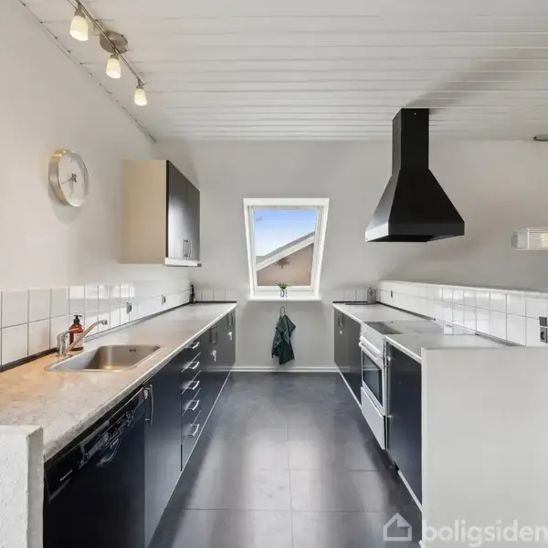Køkken med sort finish og hvide elementer. Bordplader i marmor, integreret vask, ovn med emhætte. Loftlamper og et lille vindue giver lys. Sort gulv.