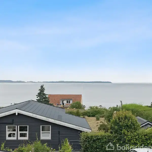 Et sort træhus står stille i forgrunden med et fladt tag. I baggrunden ses havet roligt under en blå himmel. Ingen tekst er til stede.