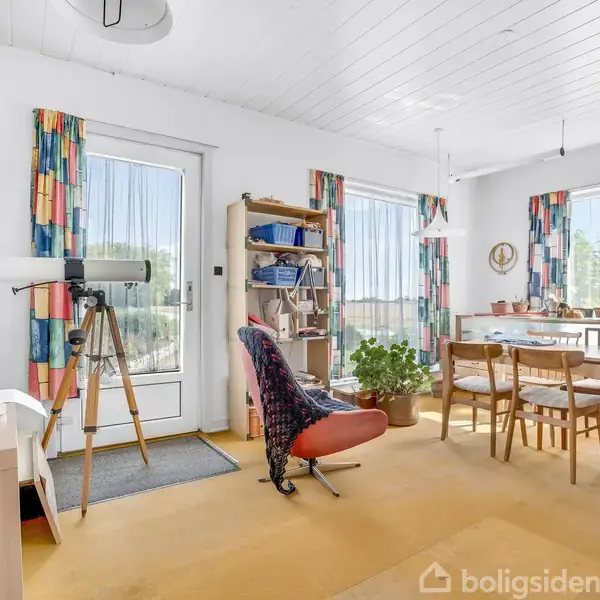 Kikkert står på stativ ved vindue bag døren med farverige gardiner, i en spisestue med bord, stole, hylde og planter. Lyse gulve og vægge skaber en lys atmosfære.