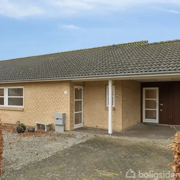 Et murstenshus med carport står i forgrunden. Huset har to vinduer, en indgangsdør, og granitskærver i indkørslen. Omkranset af hæk i et boligområde.