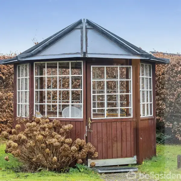 En sekskantet pavillon lavet af træ med mange vinduer står i en have. Der er en dør foran, og visne buske omgiver pavillonen. Baggrunden er en hæk.