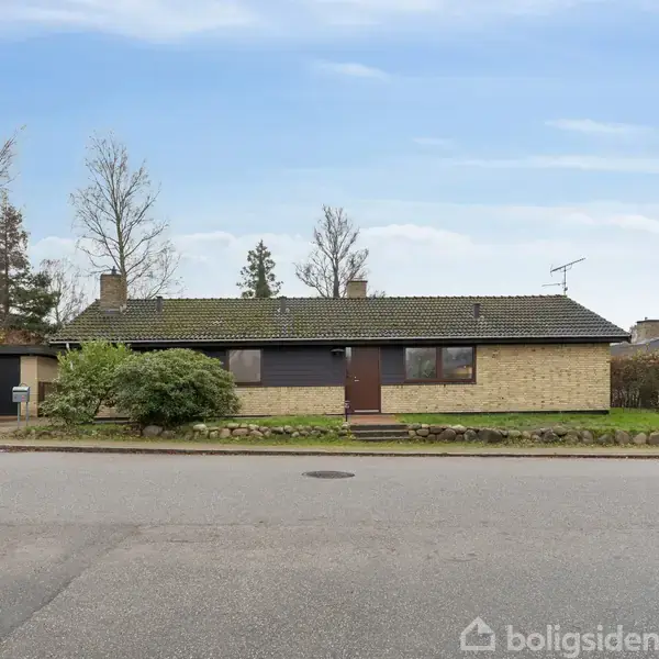 Et enfamilieshus med fladt tag og gul murstensfacade står stille omgivet af græsplæne og buske på en stille villavej.