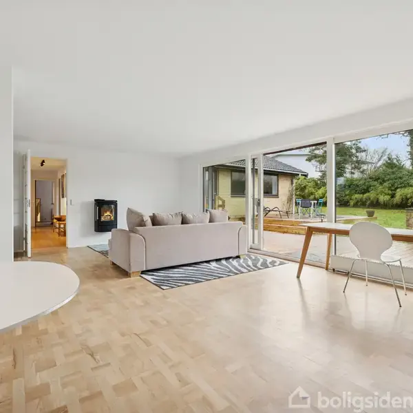 En stue med en sofa placeret på et tæppe, vendt mod en brændeovn. Store vinduer giver udsigt til en terrasse og have. En spisebord og stol er ved siden af vinduerne.