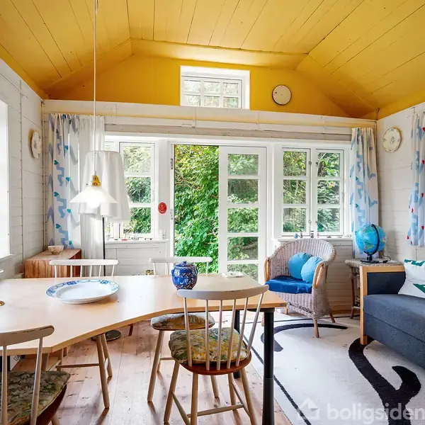 Et lyst rum med gule lofter, et spisebord, og stole. Sofa og stol står ved vinduer med udsigt til grønne buske udenfor. Globusdekoration på bord.