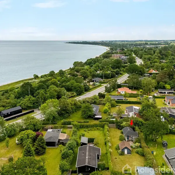 Bebyggelser ligger spredt blandt grønne områder og træer tæt på kystlinjen. En vej snor sig gennem landskabet, mens havet strækker sig i baggrunden under en klar himmel.