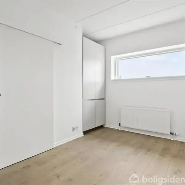 Et tomt, lyst rum med lyse træplanker på gulvet. Et smalt, rektangulært vindue over en radiator. Et indbygget skab står i hjørnet ved en skydedør.