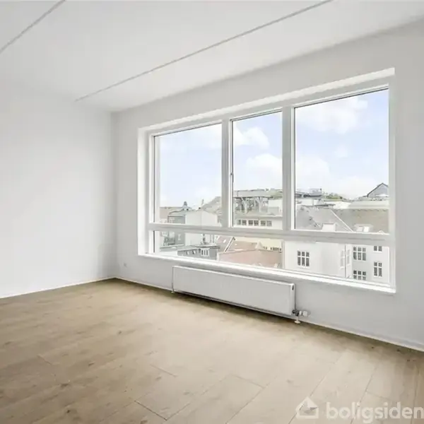 Et tomt rum har et stort vindue, der lukker lys ind. Radiator under vinduet. Hvidmalede vægge og lyst trægulv. Byudsigt i baggrunden.