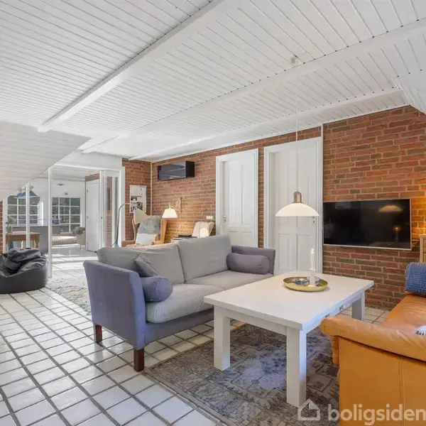 Stue med sofaer og sofabord, placeret foran en fladskærm i en hyggelig stue med skråt, hvidt loft og teglstensvægge. Der er også en pejs og flere lamper.