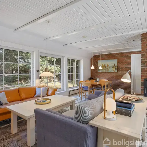 Stue med sofaer og borde omkring pejsen, placeret i et rum med store vinduer, der afslører en udendørs skovbevoksning. Rummet har hvide trævægge og gulvtæppe samt træmøbler.