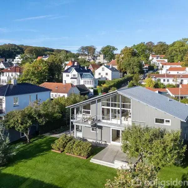 Et moderne, gråt hus med store vinduespartier står midt i en velplejet have. Omkring huset ses en række andre villaer omkranset af træer og grønne områder under en klar blå himmel.