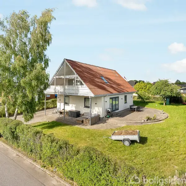 En hvid to-etagers hus med rødt tag står i en grøn have. En trailer er placeret på plænen, mens en stor træ omgiver ejendommen, der ligger i et rækkehusområde.
