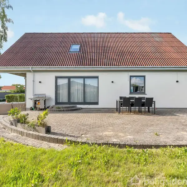 Hus med rødt tag står på en plet belagt terrasse med grill og spisebord. Græsplæne omgiver området og træer er i baggrunden under en blå himmel.