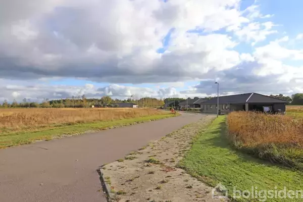 En langt, asfalteret vej løber gennem et åbent landskab med marker på begge sider. Til højre er et lavt, gråt hus ved vejen under en overskyet himmel.