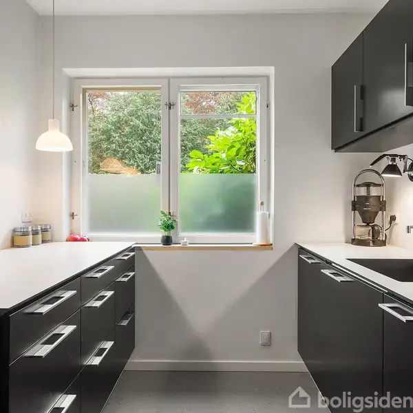 Køkken med sorte skabe og hvid bordplade, oplyst af loftslamper, står i et moderne rum. Et vindue med udsigt til grønt område bag grumset glas ses over vasken.