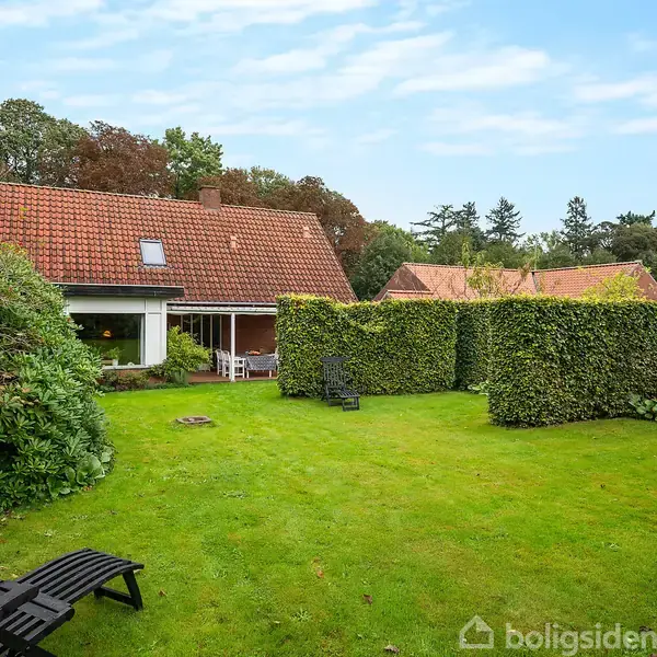 Et hus med rødt tegltag står bag en velplejet græsplæne og tætte hække. To sorte liggestole ses i haven. Omgivet af frodig natur og træer.