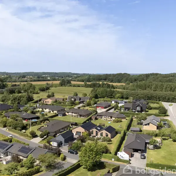Luftfoto viser et parcelhuskvarter med moderne huse omgivet af velplejede haver. Husene ligger tæt på hinanden i en grøn, naturskøn landskabsindstilling med skov og marker i baggrunden.