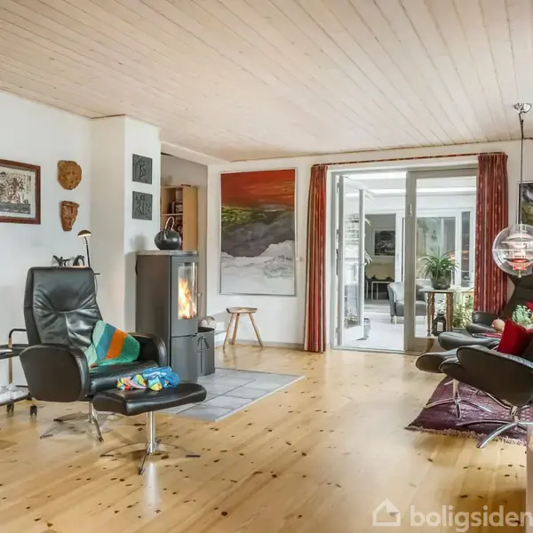 En stue med trægulv og moderne stole ved en pejs. Kunst hænger på væggene, og et rummeligt glasparti fører til et andet rum.