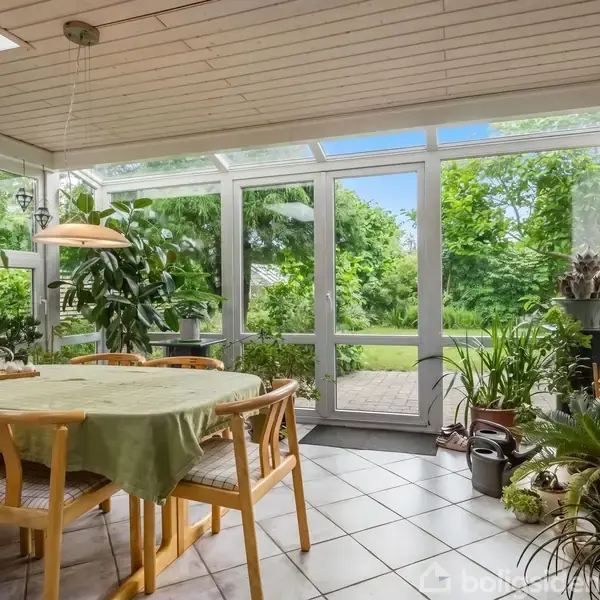 Et spisebord med seks stole står i en lys udestue, omringet af grønne planter. Store vinduer og en glasdør åbner ud til en frodig have.