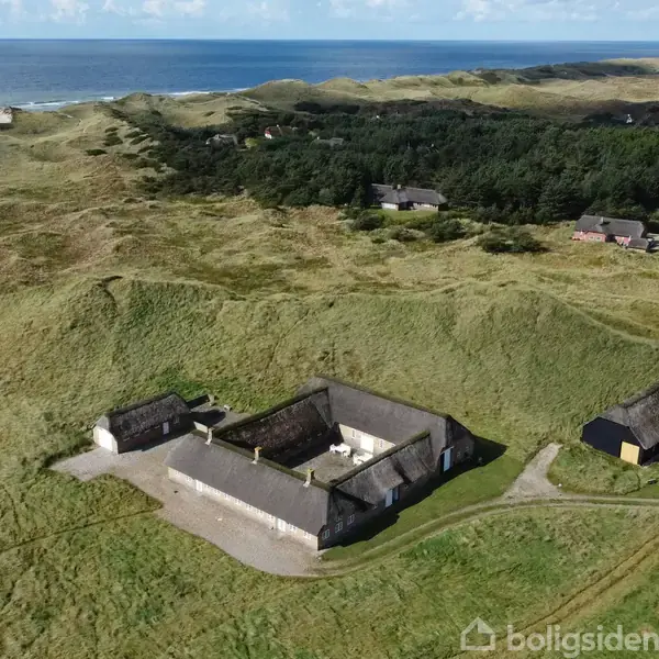 Gårdbebyggelse med stråtage står stille blandt grønne bakker. Omgivet af åbne marker og fjerne træer, med havet synligt i horisonten under en klar himmel.