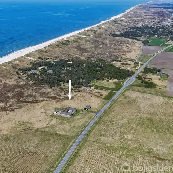 Hus ligger isoleret nær kysten, omgivet af marker og skov. En vej strækker sig langs landskabet, parallelt med havet, som ligger til venstre for billedet.