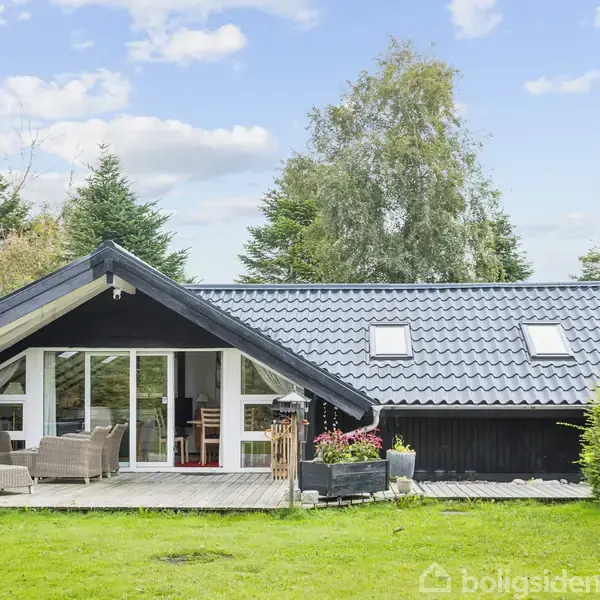 Sort træhus med store glasdøre åbne til terrasse, omgivet af grønne træer og buske under blå himmel. Møbler står på terrassen, hvilket skaber en indbydende udendørs opholdsstue.