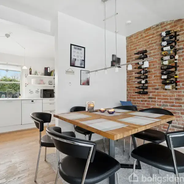 Et træbord med sorte stole rundt om står i en spisestue. En murstensvæg har vinflasker på hylder. I baggrunden ses et lyst køkken med hvide elementer og et vindue.