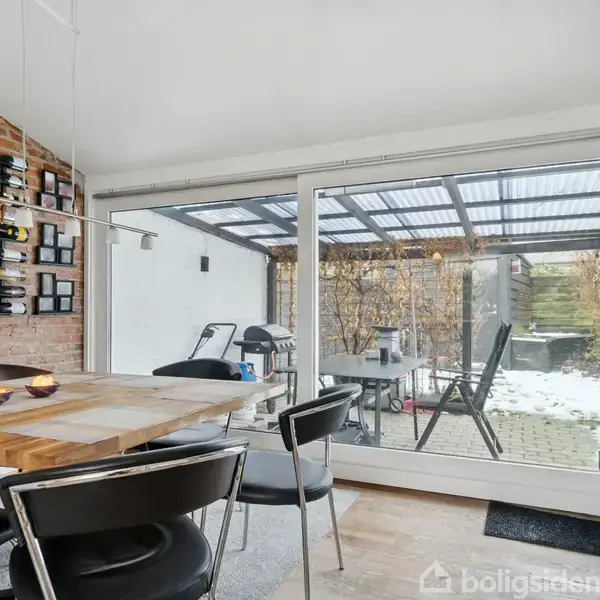 Et spisebord med sorte stole står i et moderne køkken. Udsigt gennem store glasdøre viser en overdækket terrasse med stole. Væggen har en vinkælderholder og eksponerede mursten.