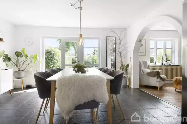 Spisebord med lammeskind og planter omkring står i en lys stue med store vinduer og en bueåbning til et sofahjørne.