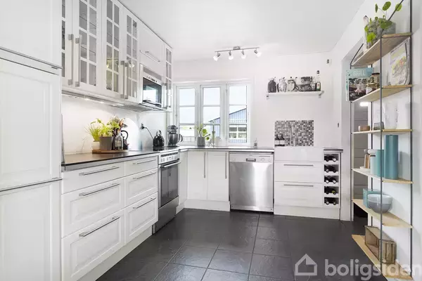 Køkken med hvide skabe og sorte fliser, der indeholder en opvaskemaskine og ovn. Hylder med nipsgenstande findes på væggen, og store vinduer giver lysindfald i rummet.