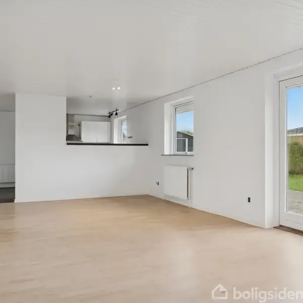 Stue med lyst trægulv, store vinduer og en havedør mod en åben græsplæne. Køkkenet ses i baggrunden, delvist skjult af en væg. Ingen tekst til stede.