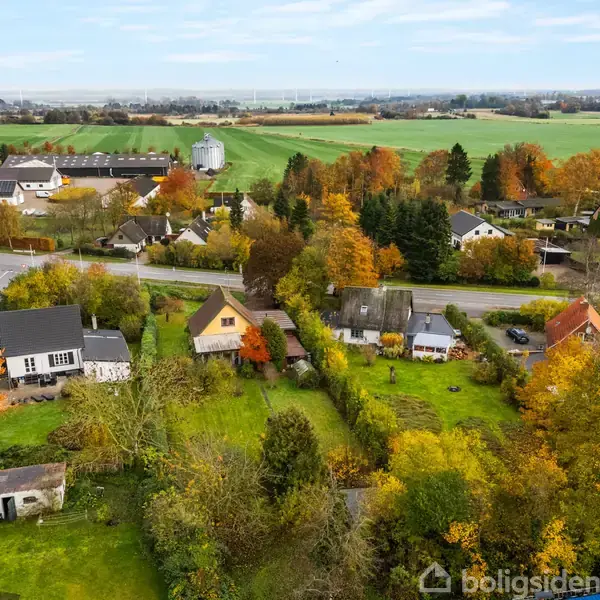 Landsbyhuse i varierede farver står stille midt i frodige, grønne områder. Omkring dem breder marker sig ud i horisonten, og træerne viser efterårets gyldne nuancer.