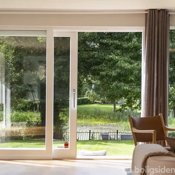 Glasdør står åben til en terrasse, indrammet af gardiner. Indenfor ses en stue med lænestol, sofa og et bord med en plante. Udenfor er en grøn have.
