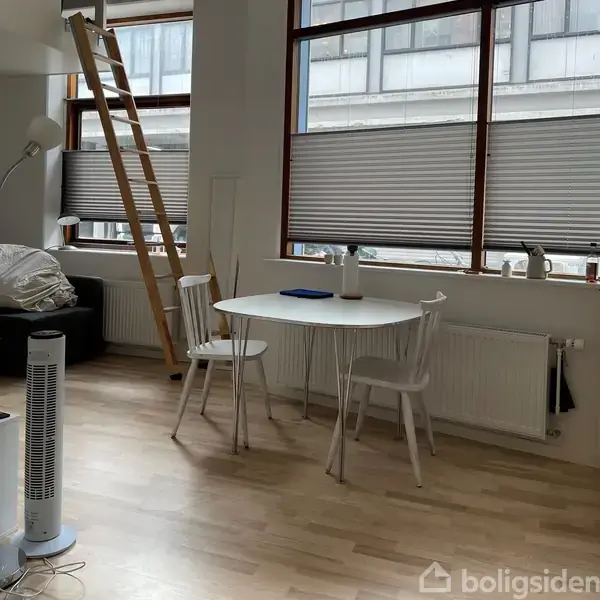 Et rundt, hvidt bord med to stole står ved siden af et vindue med skodder. En stige leder op til en hems. Lampe og sofa ses i baggrunden. Inget tekst er fundet.
