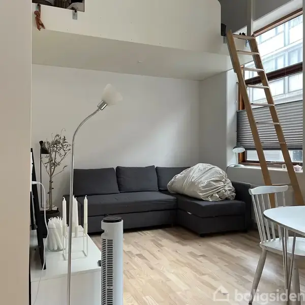 Grå sofa står i hjørnet, dækket med et hvidt tæppe. En lampe ved siden af lyser op. Stige fører til en hems ved vinduet i en moderne stue.