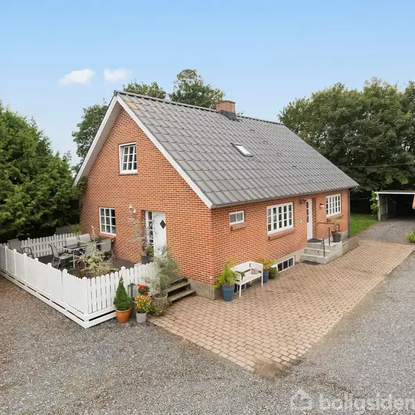 Rødstenshus med gråtag står stille på en gårdsplads belagt med småsten. Ved siden af huset er et hvidt hegn omgivet af planter. I baggrunden ses tættere beplantning.