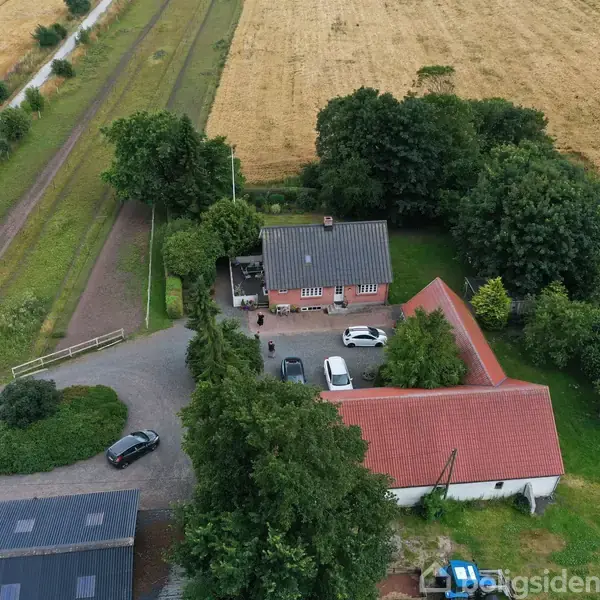 Et rødt murstenshus omgivet af en gårdsplads med parkerede biler, træer og marker. Beliggende ved siden af en grusvej og store markarealer.